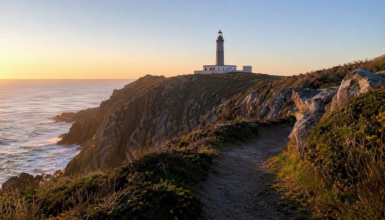 El faro de Corrubedo se alza majestuoso sobre los acantilados, bañados por los últimos rayos del sol al atardecer.